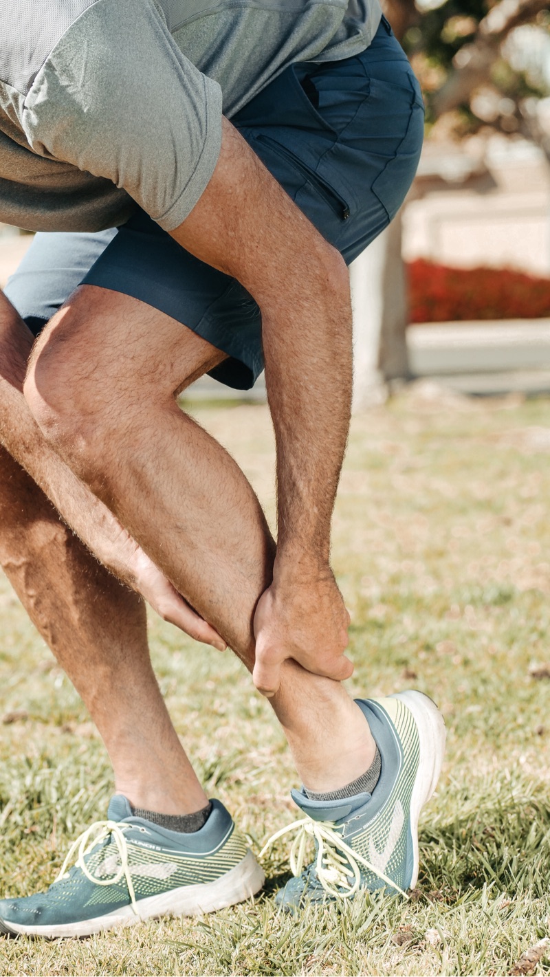 Man in pain reaching for his ankle while wearing running shoes outdoors